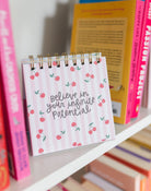 Small spiral-bound notebook with 'Believe in your infinite potential' text on a shelf with books.