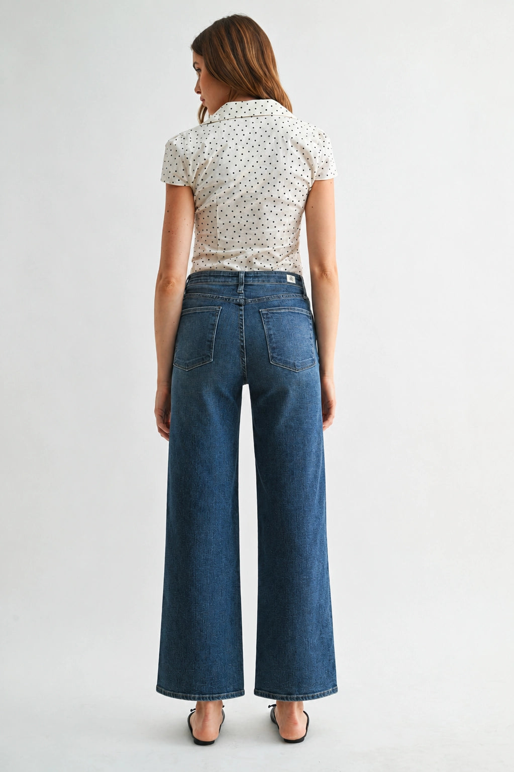 A woman shown from the back wearing a cream short-sleeve blouse with a small black polka dot pattern and high-waisted, wide-leg blue denim jeans, set against a plain light gray studio background.