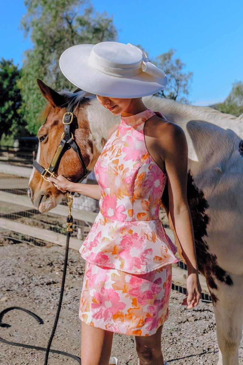 Woman in a floral dress and white hat standing next to a horse in an outdoor setting.