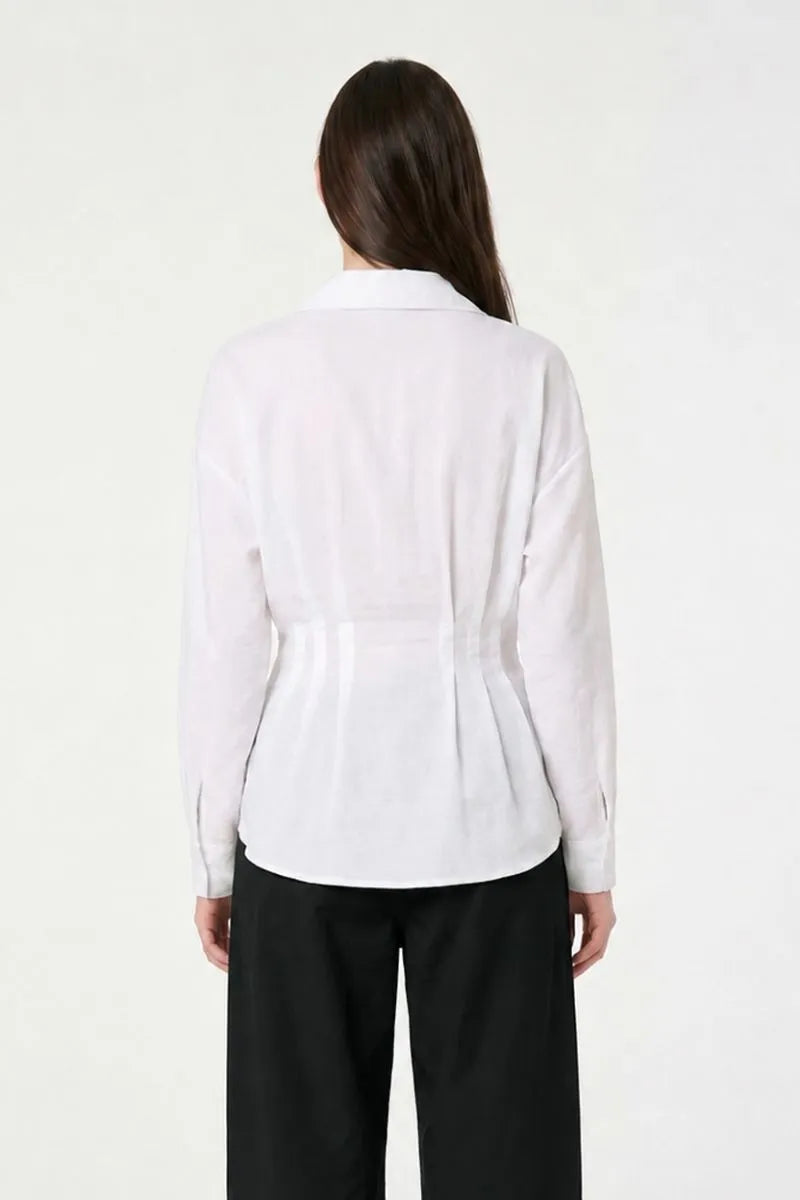 A woman viewed from the back wearing a white long-sleeve collared blouse with a gathered waist seam and black wide-leg pants, against a plain light background.