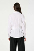 A woman viewed from the back wearing a white long-sleeve collared blouse with a gathered waist seam and black wide-leg pants, against a plain light background.