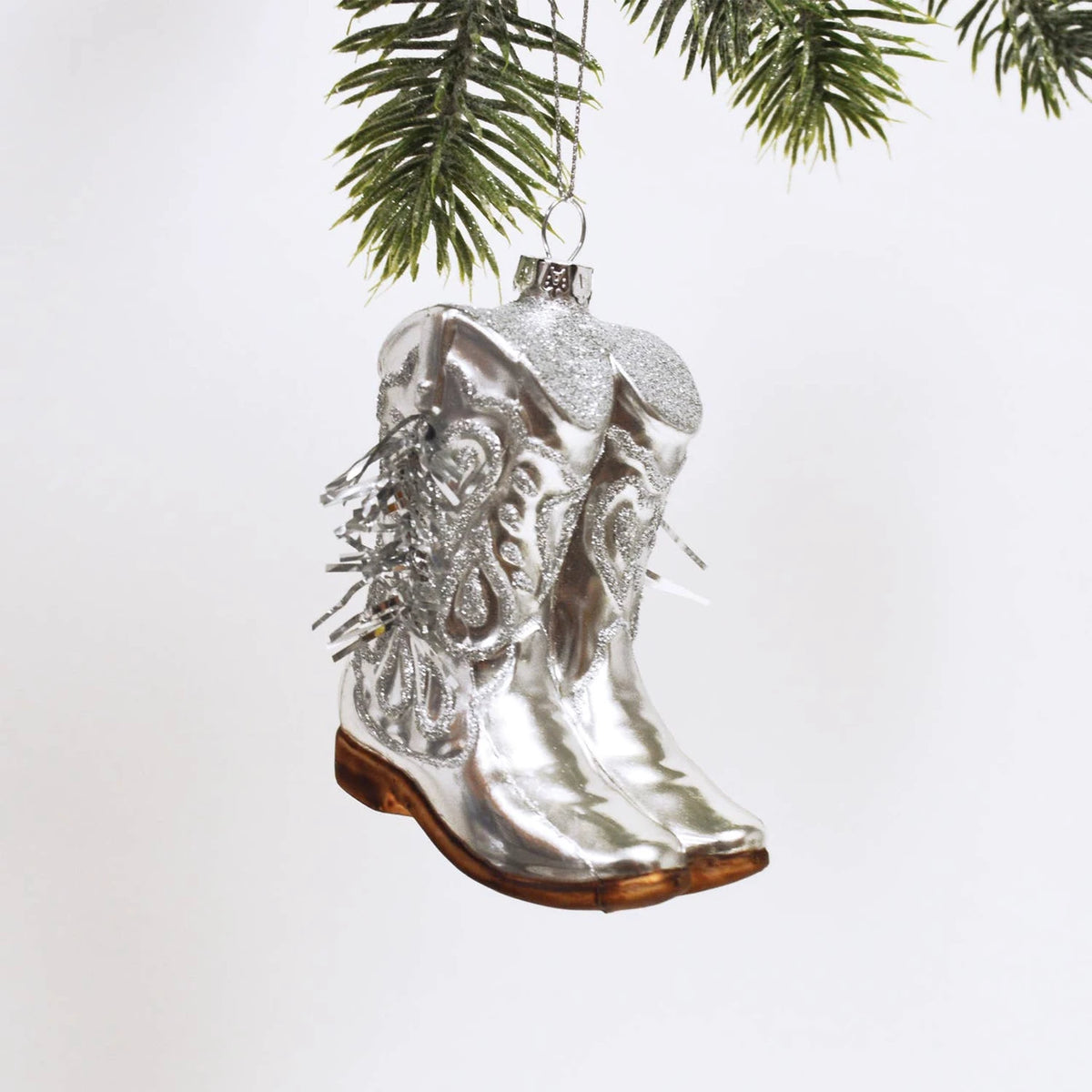 Silver cowboy boot ornament hanging from a tree branch on a white background