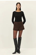 A model stands against a plain light background wearing a fitted black long-sleeve top, brown shorts, sheer black tights, and black knee-high boots.