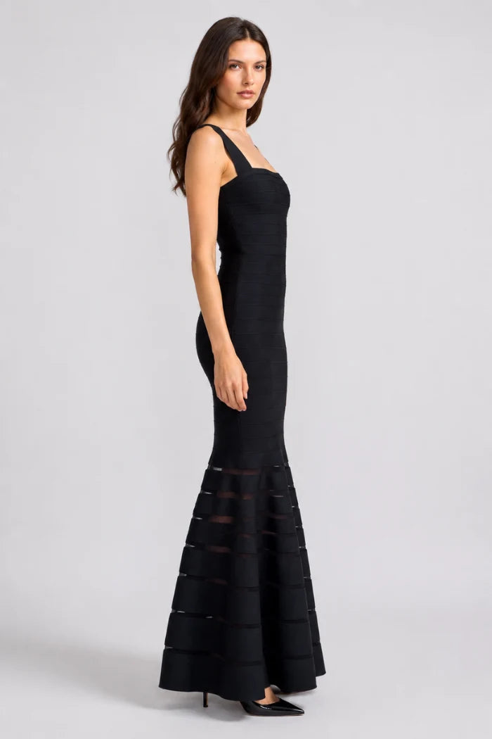 A woman modeling a black sleeveless floor-length gown with a fitted bodice and a flared skirt featuring sheer horizontal bands near the hem, photographed against a light gray studio background.