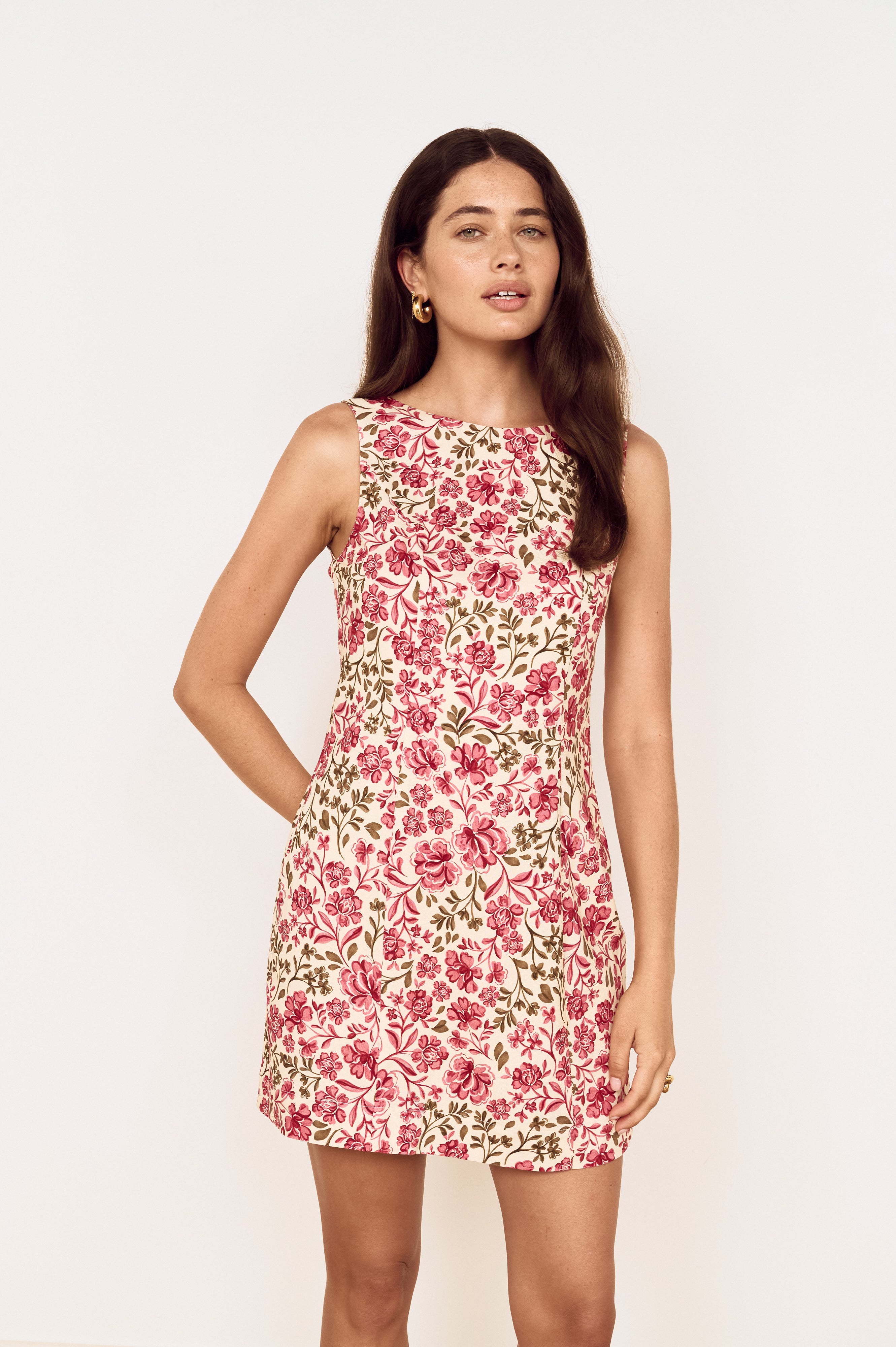 A woman modeling a sleeveless fit-and-flare dress with a pink and red floral print on a cream background, set against a plain off-white studio backdrop.