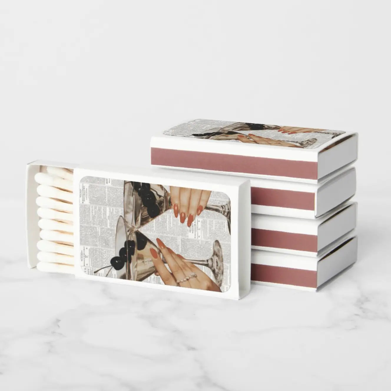 A set of decorative matchboxes with a vintage-style collage print featuring hands, nails, and a glass, displayed on a light marble surface against a white background.