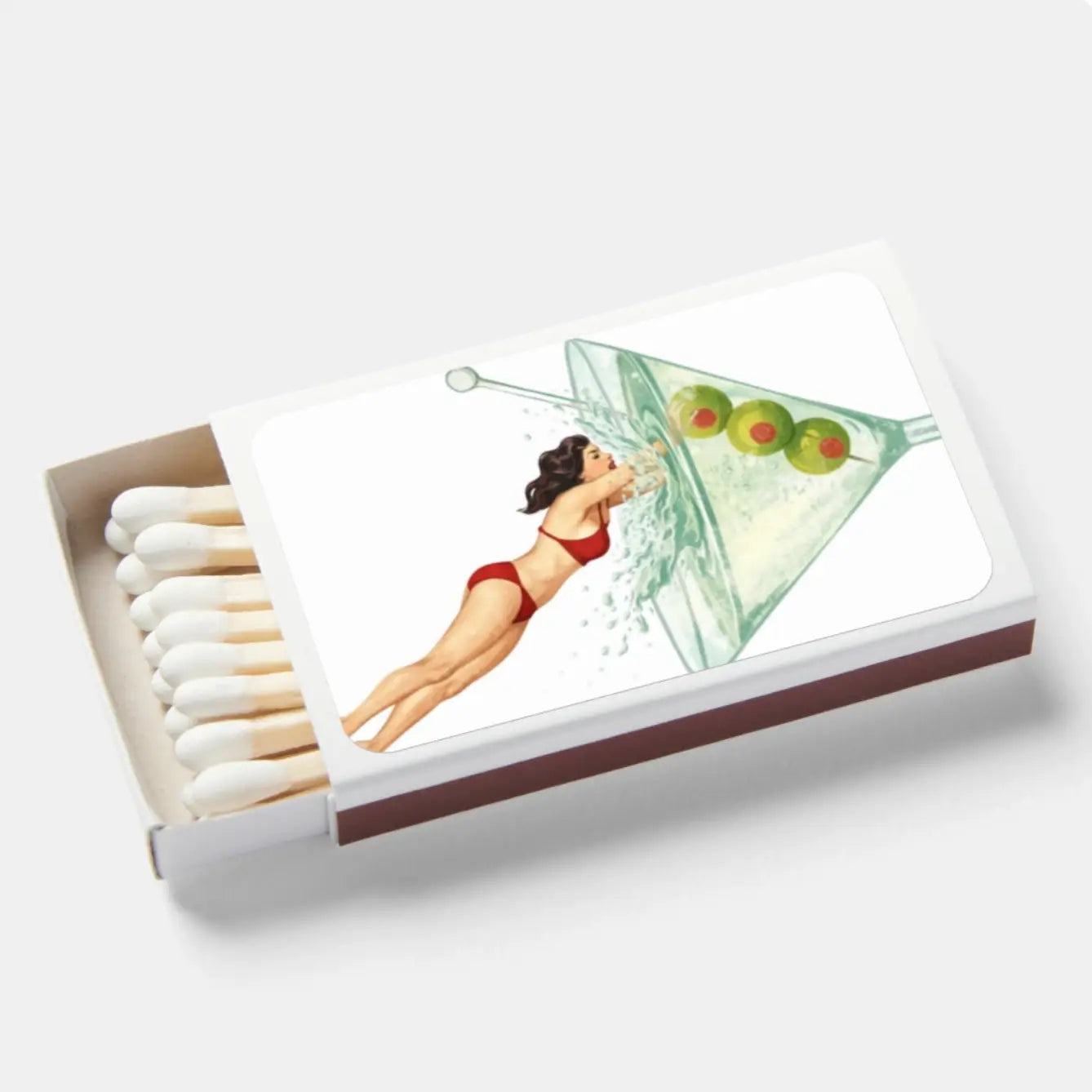 A novelty matchbox with a white lid featuring a retro illustration of a woman in a red bikini reaching into a martini glass with olives, and white matches visible in the open box.