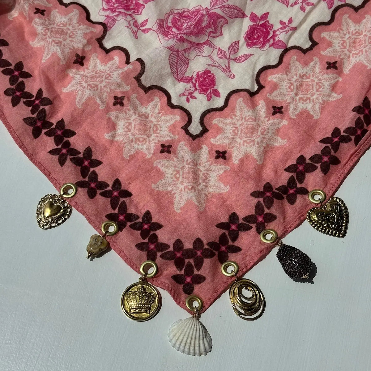 A pink and white printed scarf with dark brown floral embroidery along the border, decorated with hanging gold-tone charms including a shell, a small bead tassel, and metallic medallions, laid flat on a light background.