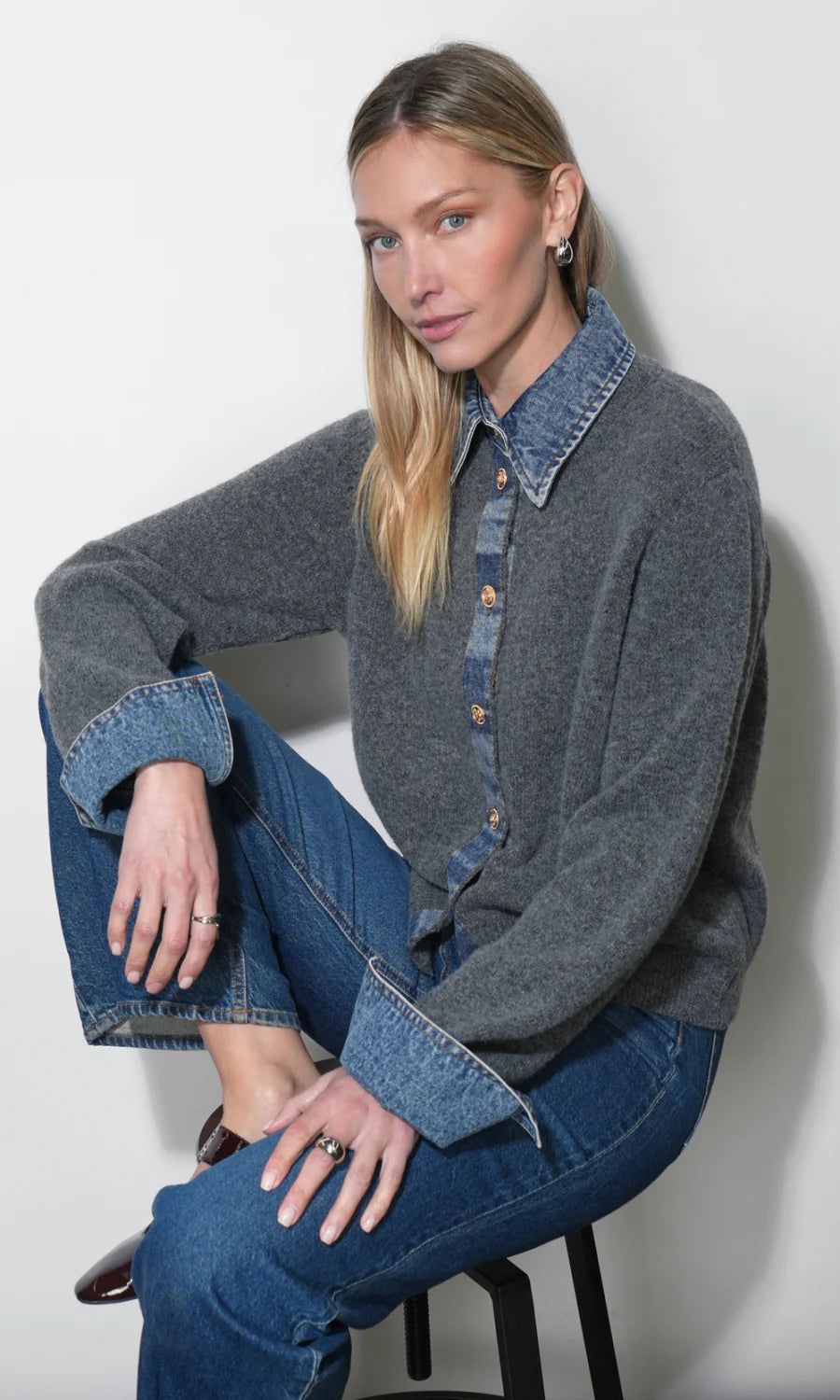 A woman sits on a stool wearing a charcoal gray knit button-up cardigan with contrasting blue denim collar and cuffs, paired with blue jeans, against a plain light gray wall.