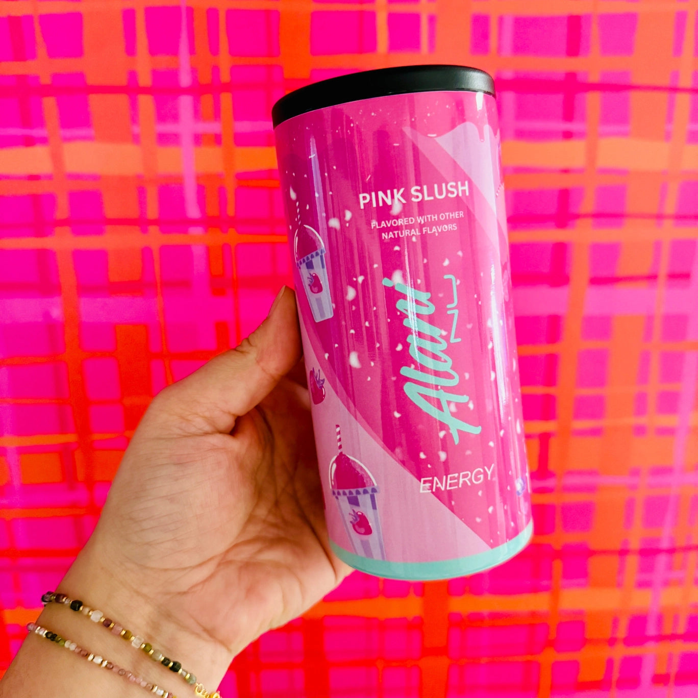 Pink energy drink can held by a hand against a colorful plaid background