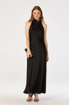A woman modeling a sleeveless black satin halter maxi dress with a draped cowl neckline, standing against a plain white studio background and holding a small black clutch.