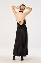 A woman viewed from the back wearing a black satin halter maxi dress with a tie-back detail and black heels, set against a plain light gray studio background.