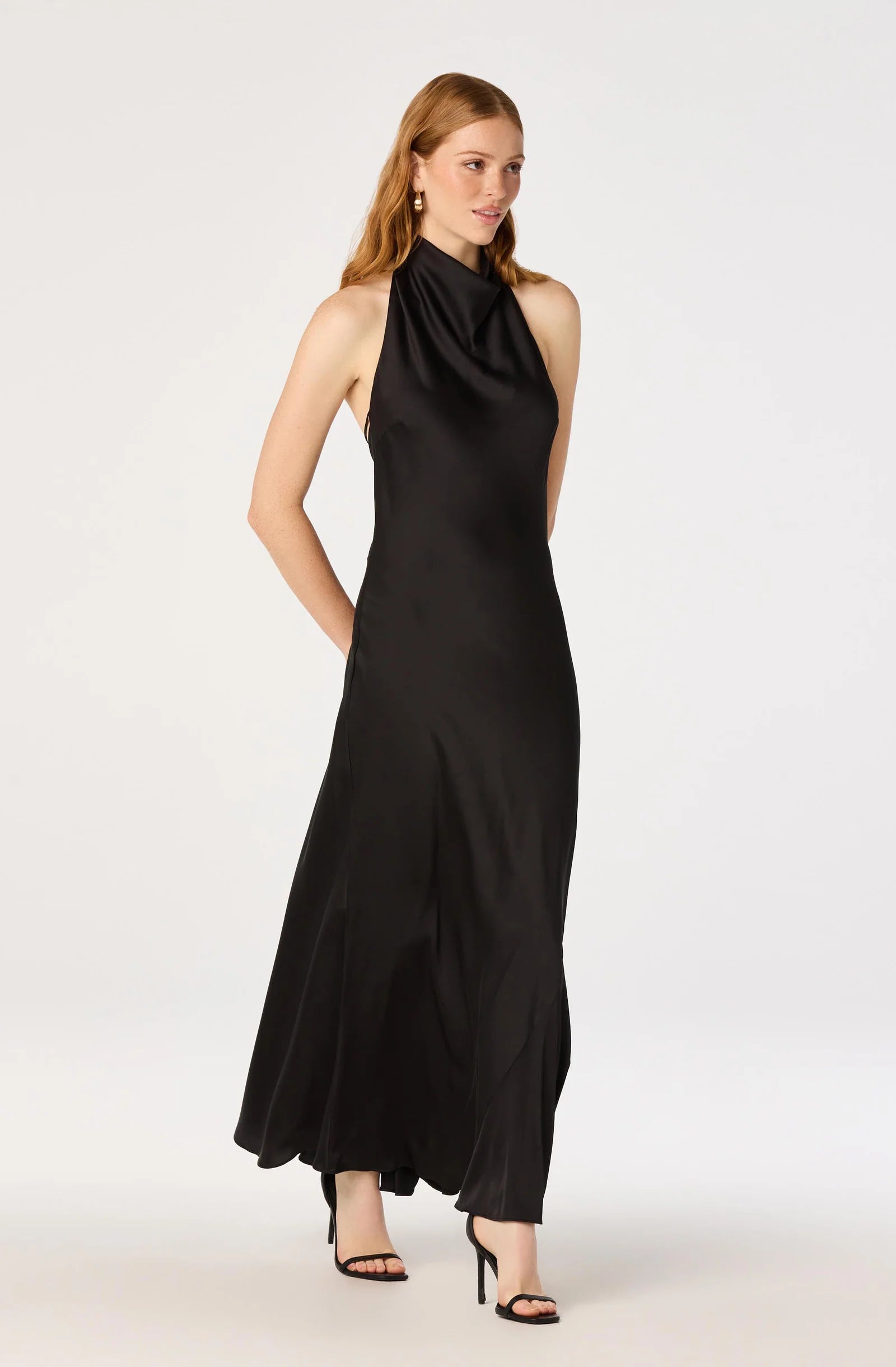 A woman modeling a sleeveless black halter-neck maxi dress with a draped satin-like finish, paired with black high-heeled sandals, against a plain light gray studio background.