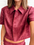 A close-up of a model wearing a short-sleeve collared crop top in a dusty rose or mauve suede-like fabric, paired with matching bottoms, set against a white brick wall.