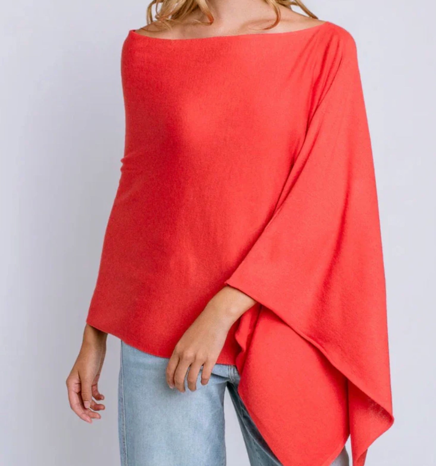 A woman wearing a bright coral-red off-the-shoulder poncho top with light blue jeans, shown from the neck down against a plain light gray background.