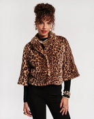 A woman modeling a cropped leopard-print faux fur jacket with wide short sleeves, paired with black pants and jewelry, against a plain light gray background.