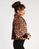 A woman in profile wearing a leopard-print faux fur jacket over a black long-sleeve top and black pants, photographed against a clean white studio background.
