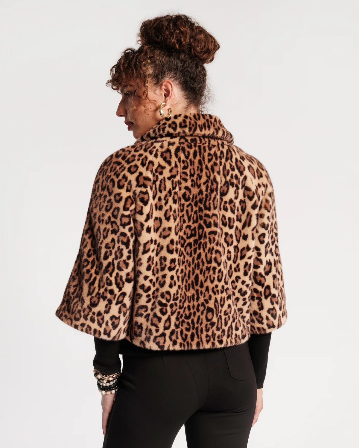 A woman shown from the back wearing a leopard-print faux fur cape jacket over a black top and black pants, against a plain white studio background.
