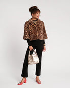 A woman modeling a cropped leopard-print faux fur jacket over a black long-sleeve top and black pants, holding a metallic rose gold handbag and wearing red heels, against a plain white studio background.