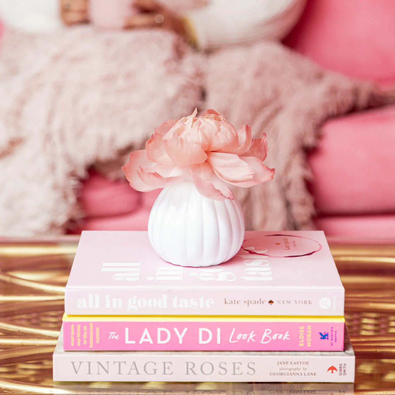 Stack of books titled 'The Lady Di Look Book' and 'Vintage Roses' with a small white vase and pink flower on top, against a soft pink background.