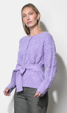 A woman modeling a lavender textured knit cardigan with a tie waist belt, paired with gray trousers, against a plain off-white wall.