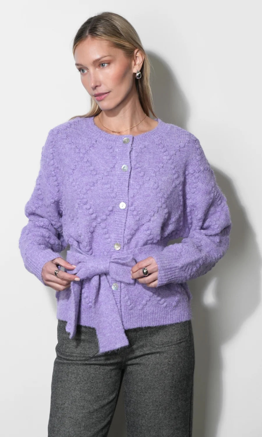 A woman wearing a lavender textured knit cardigan with a button front and self-tie belt, paired with dark gray trousers, standing against a plain off-white wall.