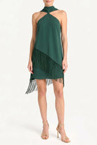 A woman models a dark green halter-neck dress with a diagonal fringe detail, paired with gold high-heeled sandals against a plain white studio background.