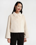 A woman modeling a cropped, cream-colored fuzzy button-up jacket with a collar and wide sleeves, paired with black pants, against a plain light gray background.