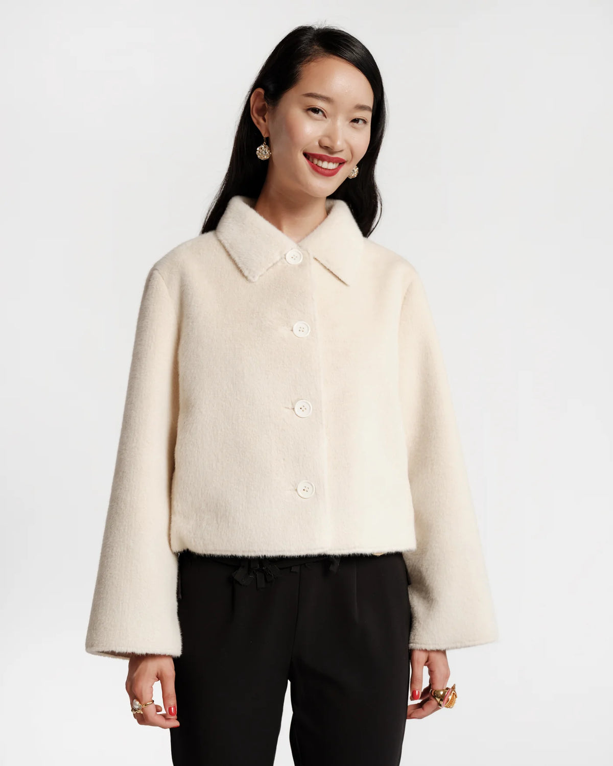 A woman modeling a cropped, cream-colored fuzzy button-up jacket with a collar and wide sleeves, paired with black pants, against a plain light gray background.