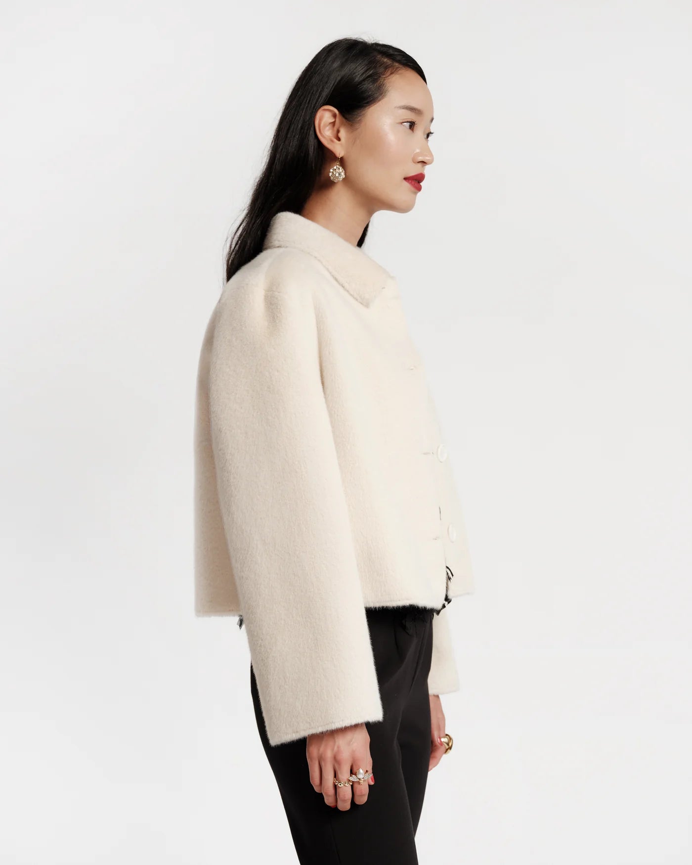 A woman shown in profile wearing a cropped, long-sleeve cream-colored textured jacket with black pants, set against a plain light gray studio background.