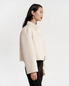 A woman shown in profile wearing a cropped, long-sleeve cream-colored textured jacket with black pants, set against a plain light gray studio background.