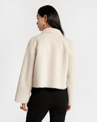 A woman shown from the back wearing a cropped, long-sleeve cream-colored fuzzy jacket and black pants against a plain white studio background.