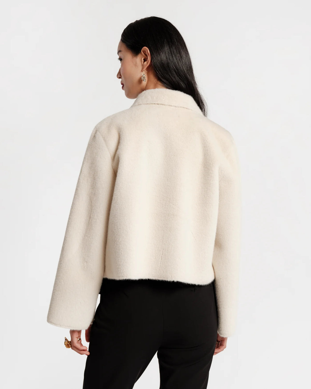 A woman shown from the back wearing a cropped, long-sleeve cream-colored fuzzy jacket and black pants against a plain white studio background.