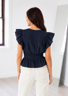 Woman wearing a navy blue top with ruffled sleeves in a minimal white room.