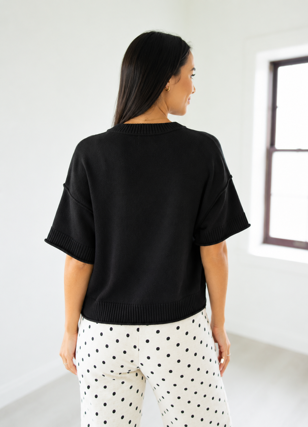 Woman wearing a black sweater and polka dot pants in a minimal white room.
