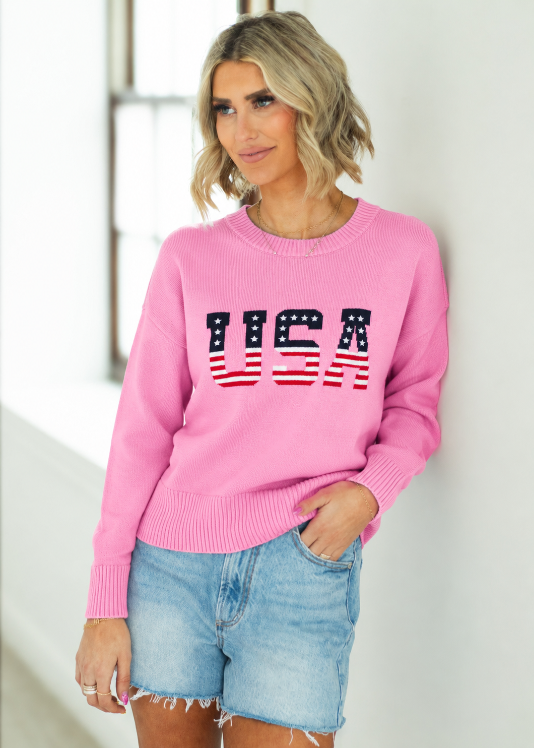 Woman wearing a pink sweater with 'USA' print and denim shorts against a white wall.