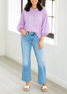 Woman wearing a light purple shirt and blue jeans standing in a bright room with large windows.