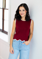 Woman wearing a red sleeveless top and light blue jeans against a white wall.