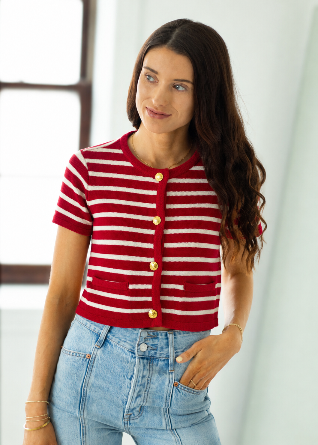 Woman wearing a red and white striped cardigan with gold buttons, standing against a white curtain.