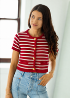 Woman wearing a red and white striped cardigan with gold buttons, standing against a white curtain.
