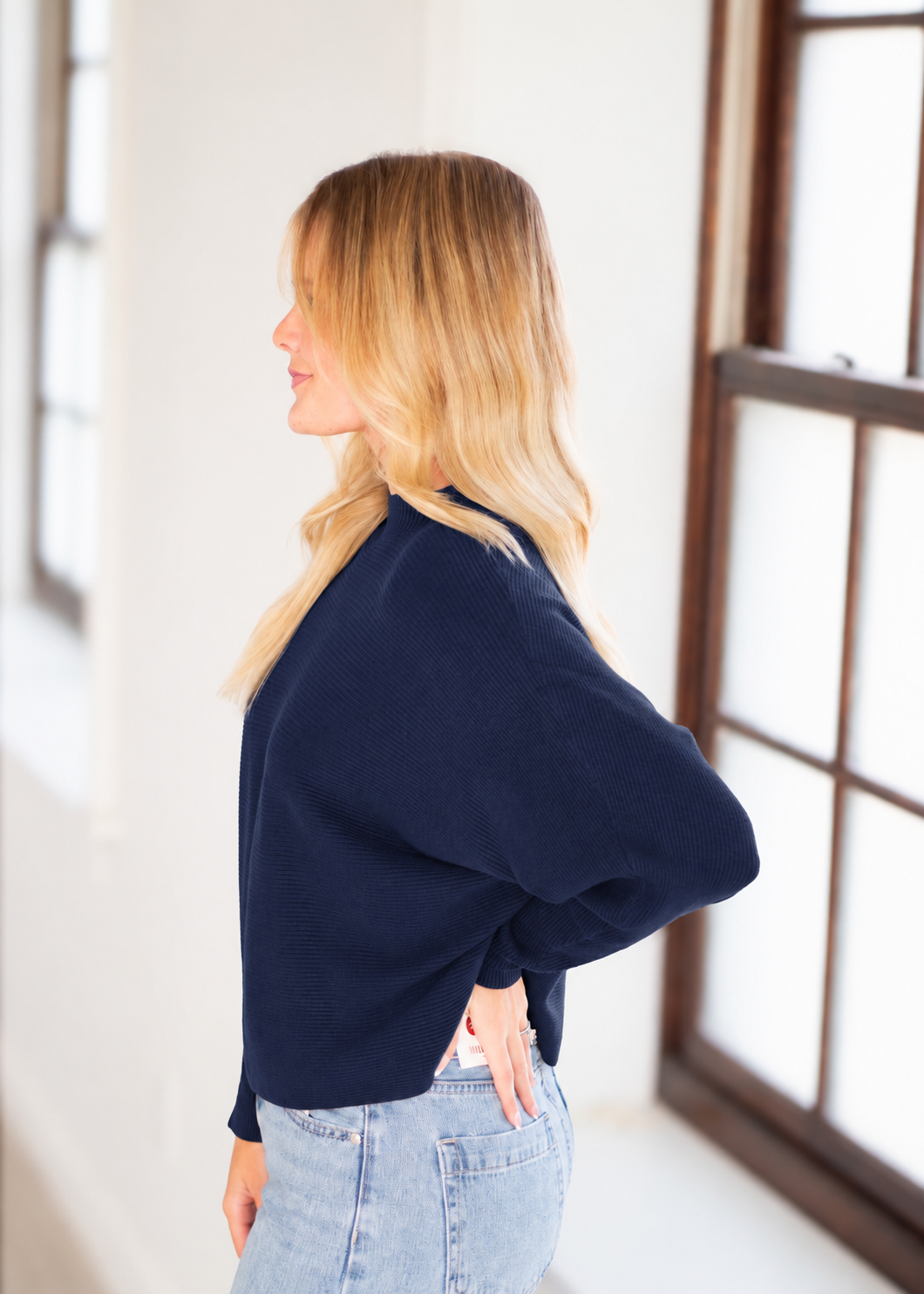 A woman with blonde hair is shown from the side profile wearing a navy blue long-sleeve top and light blue jeans, standing in a brightly lit indoor space with large windows.