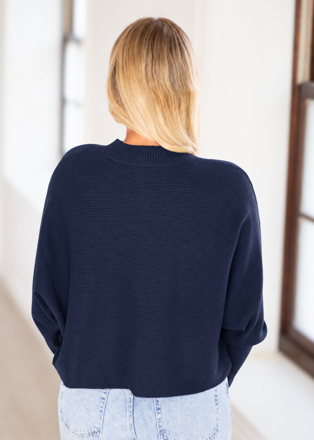 A woman viewed from behind wearing a dark navy blue ribbed knit sweater and light wash denim jeans in a bright indoor setting.