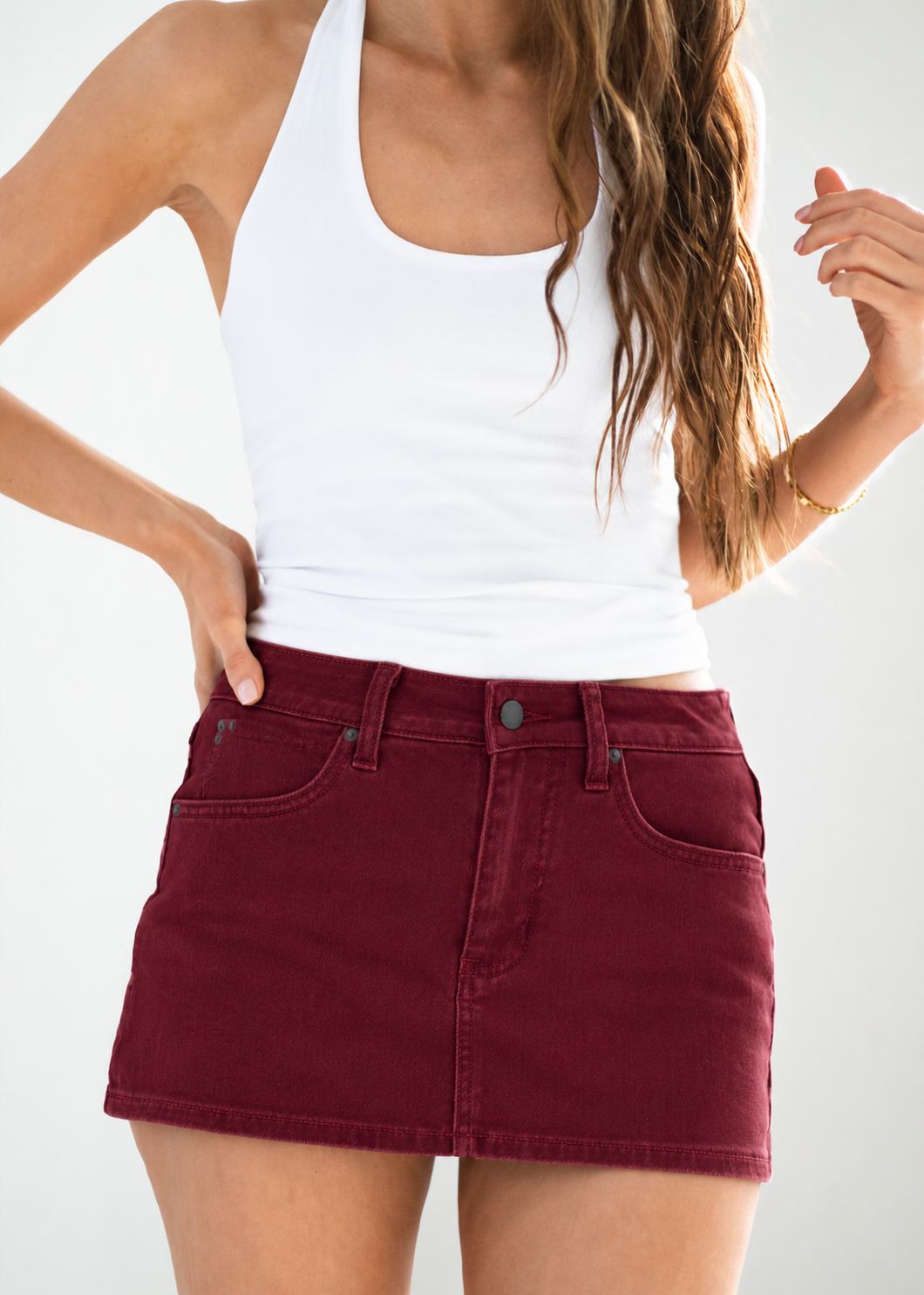 A woman wearing a white ribbed tank top and a high-waisted, dark burgundy denim mini skirt with front patch pockets, shown from the chest down against a light gray studio background.