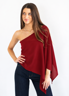 A woman modeling a deep red one-shoulder draped top with dark blue jeans, standing against a plain white background.
