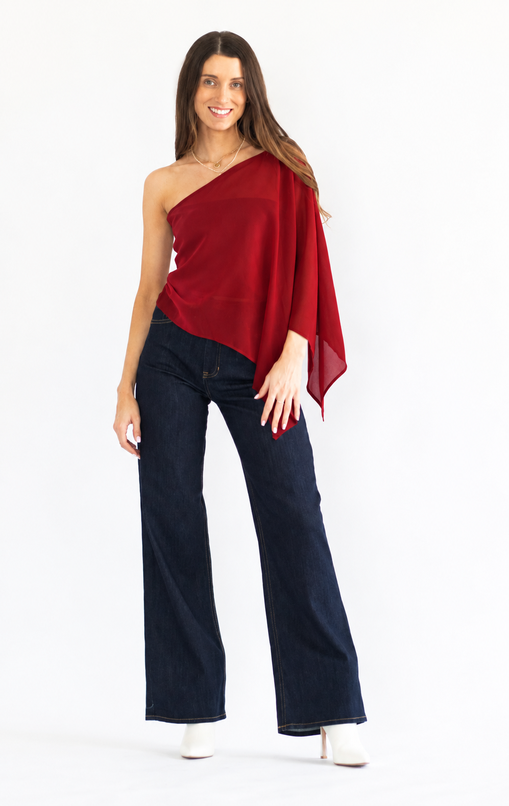 A woman modeling a dark red one-shoulder draped top paired with dark blue wide-leg jeans and white heeled shoes, photographed against a plain white studio background.