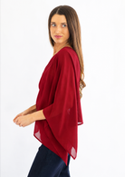 A woman wearing a red draped, semi-sheer three-quarter sleeve top paired with dark blue pants, photographed against a solid white background.
