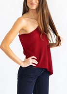 A woman modeling a one-shoulder red knit top with dark blue jeans, standing against a plain white studio background.