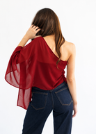 A woman shown from behind wearing a deep red one-shoulder sheer blouse with wide flowing sleeves and dark blue jeans, set against a plain white background.