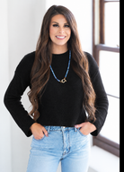 A woman with long brown hair wearing a black ribbed knit long-sleeve top and light blue jeans, accessorized with a blue beaded necklace with a gold clasp, standing indoors near a window.