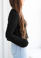 A woman shown from the side wearing a black textured knit long-sleeve sweater and light blue jeans against a bright white background.